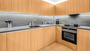 A modern kitchen with wooden cabinets, a stainless steel sink, a built-in oven, and a black countertop. - AC | Private Balcony | Gym  | StayCozy (London)