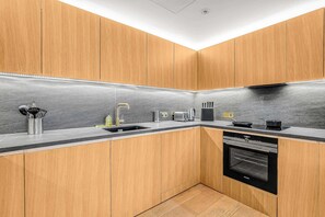 A modern kitchen with wooden cabinets, a stainless steel sink, a built-in oven, and a black countertop. - AC | Private Balcony | Gym  | StayCozy (London)