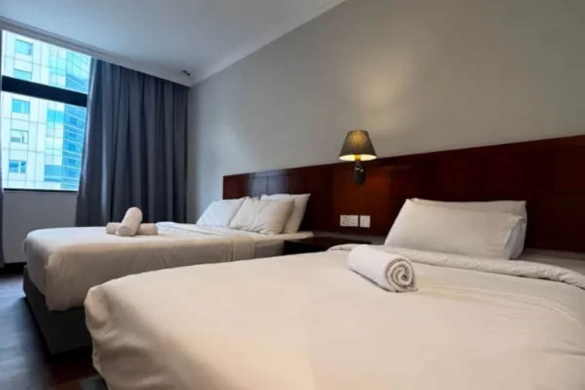 A hotel room with two beds, a window with curtains, a wall-mounted lamp, and a view of a building outside.