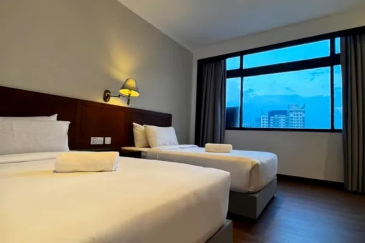 A hotel room with two beds, a nightstand, a lamp, and a large window with a view of buildings.