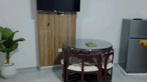 Een kamer met een houten kast, een flatscreen televisie, een klein rond tafeltje met stoelen, een potplant en een koelkast.