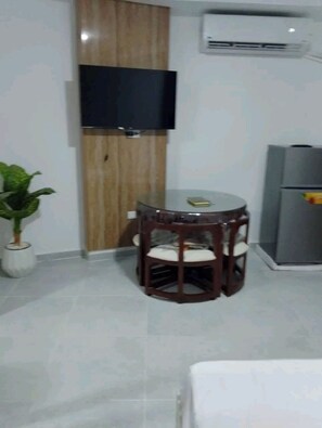 Ein Zimmer mit einem Holzkasten, einem Flachbildfernseher, einem kleinen runden Tisch mit Stühlen, einer Zimmerpflanze und einem Kühlschrank.