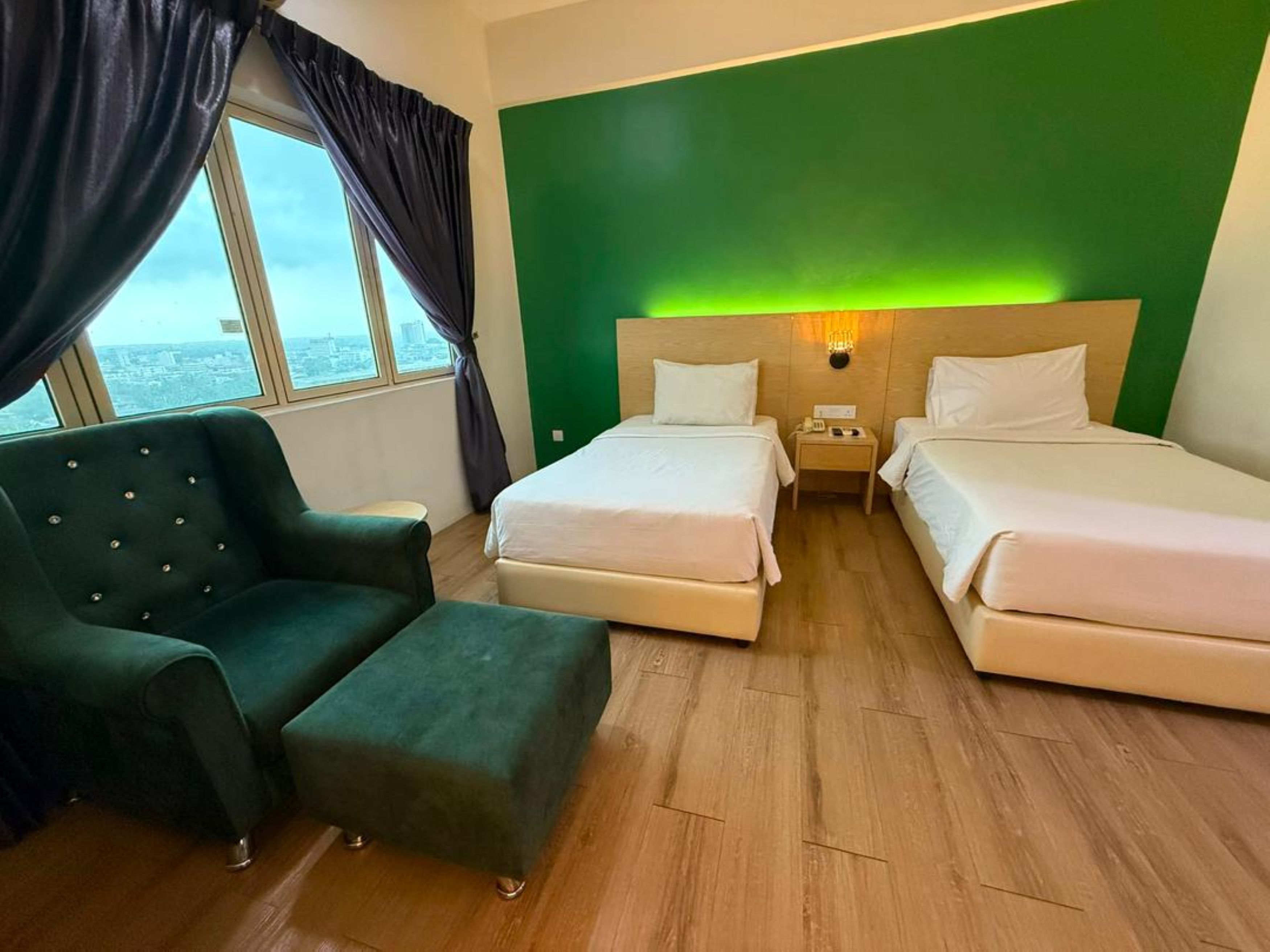 A hotel room with two beds, a green wall, a view through a window, and a green armchair.