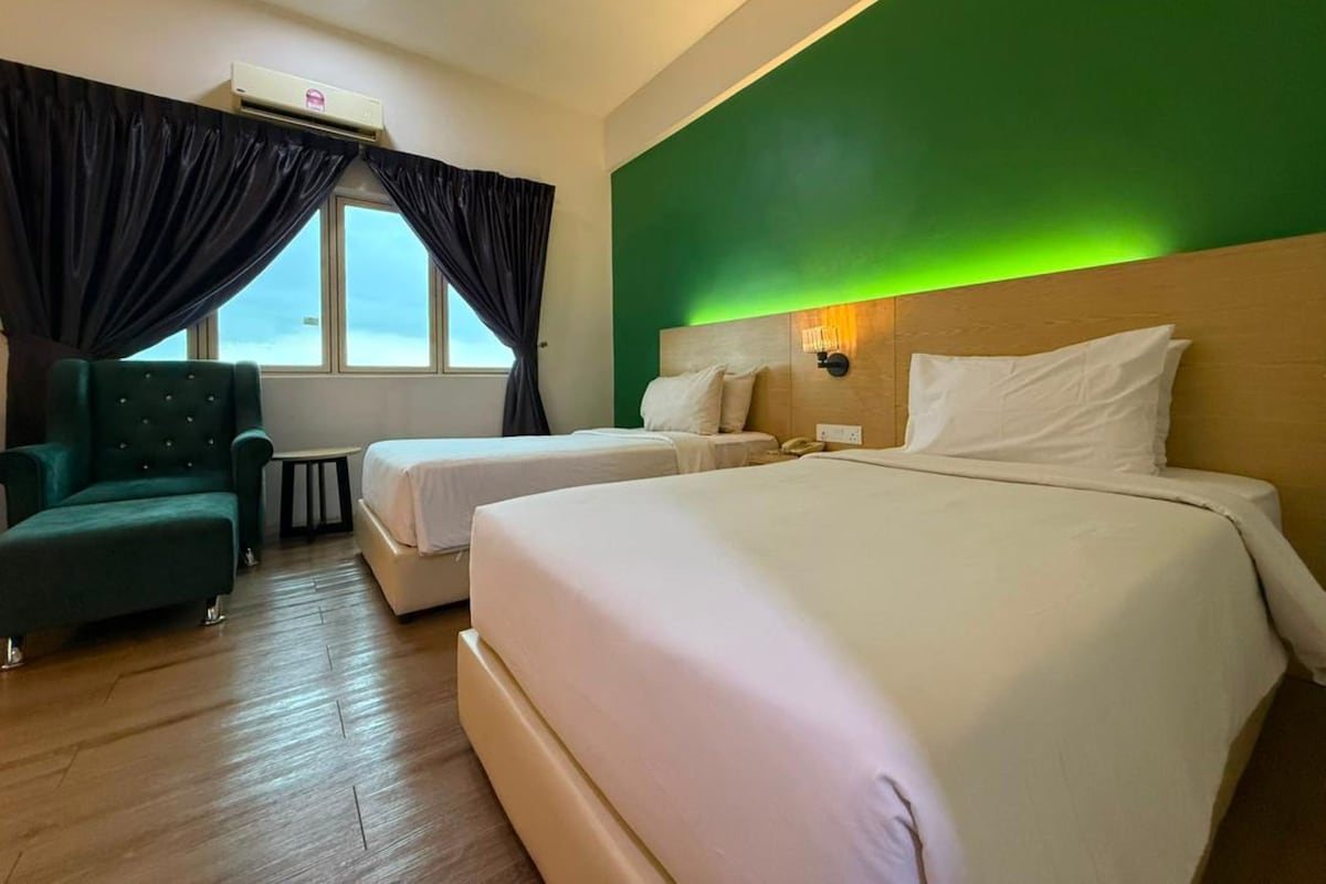 Habitación de hotel con dos camas, una pared verde, una ventana con cortinas, un sillón verde y una mesita de noche.