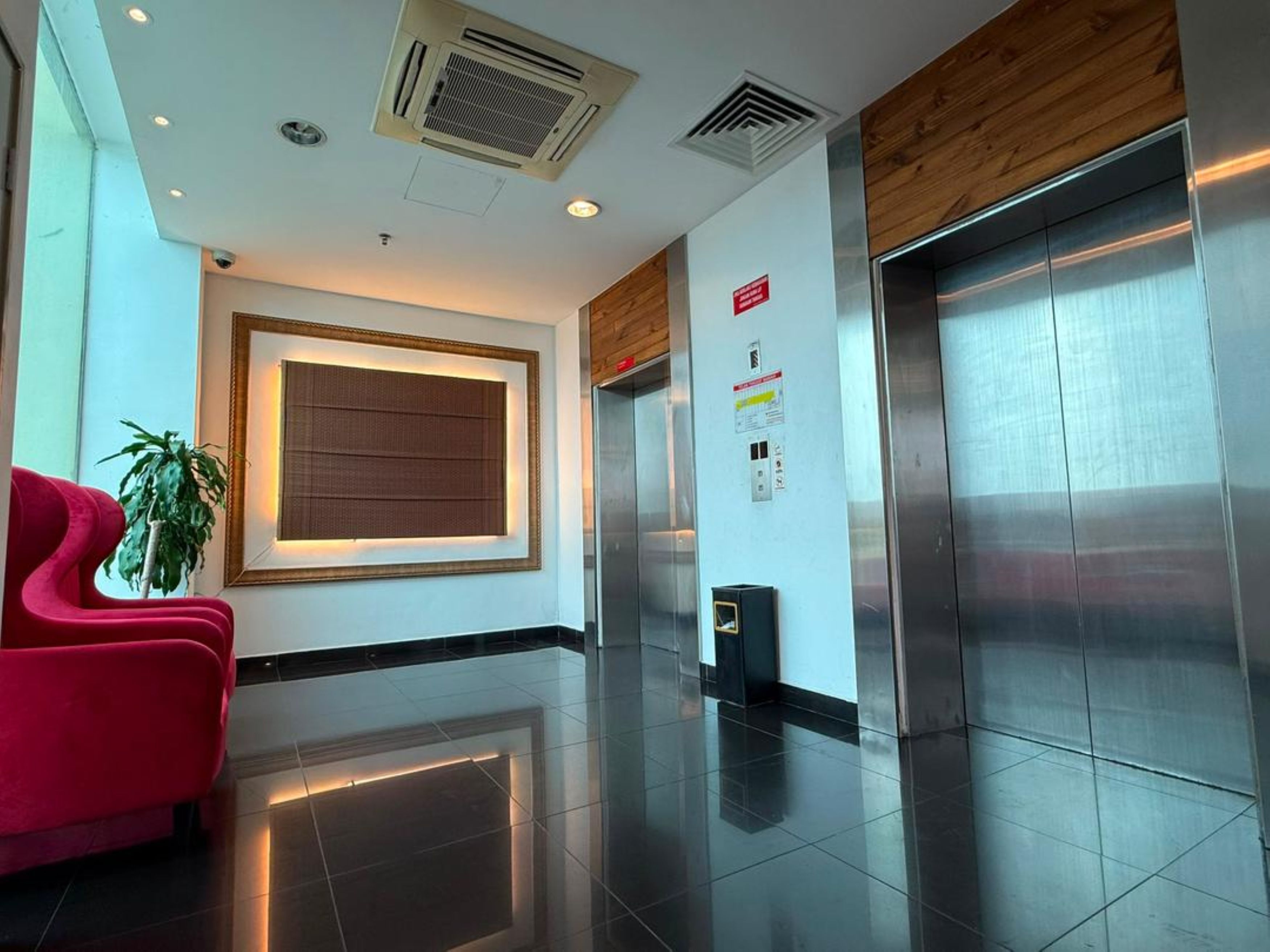 A modern lobby with a red chair, a potted plant, a large wall-mounted screen, and an elevator.