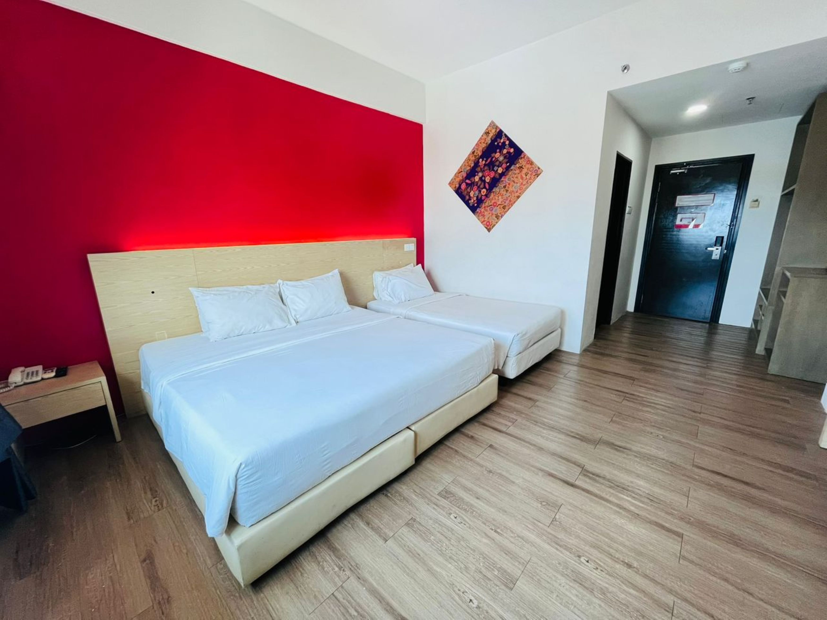 Habitació d'hotel amb un llit gran, una taula de nit, una paret d'accent vermella i una porta que dona a un passadís.