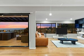 A modern living room with a sofa, a TV, and a dining area with a table and chairs. - Terrigal Beach House | 180° Sea View, Private Pool (Terrigal)