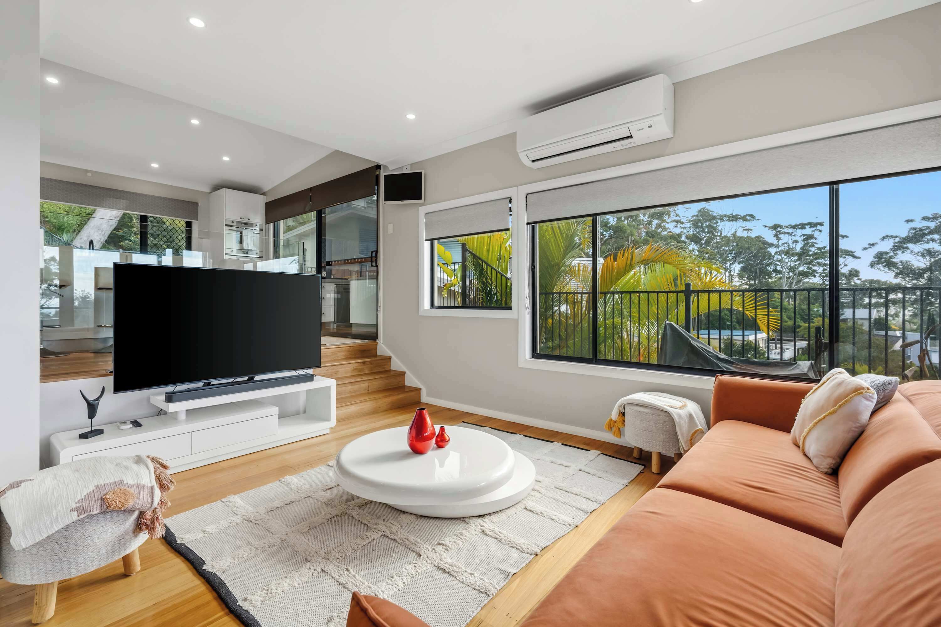 A modern living room with a flat-screen TV, a sofa, a coffee table, and a large window with a view of greenery.