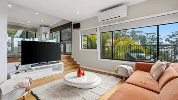 A modern living room with a flat-screen TV, a sofa, a coffee table, and a large window with a view of greenery.