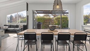 Un comedor moderno con una mesa grande y sillas, una lámpara colgante y ventanas amplias que ofrecen vistas al exterior.