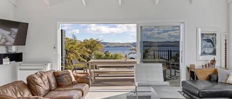 A modern living room with a leather sofa, a white armchair, and a coffee table. There is a large window offering a view of the sea and hills.