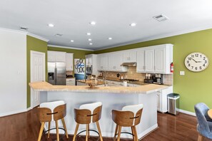 A modern kitchen with white cabinets, a central island with bar stools, a clock on the wall, and a stainless steel refrigerator. - Ultimate family friendly retreat: pool, spa & epic game-room in Frisco, TX. (Frisco)