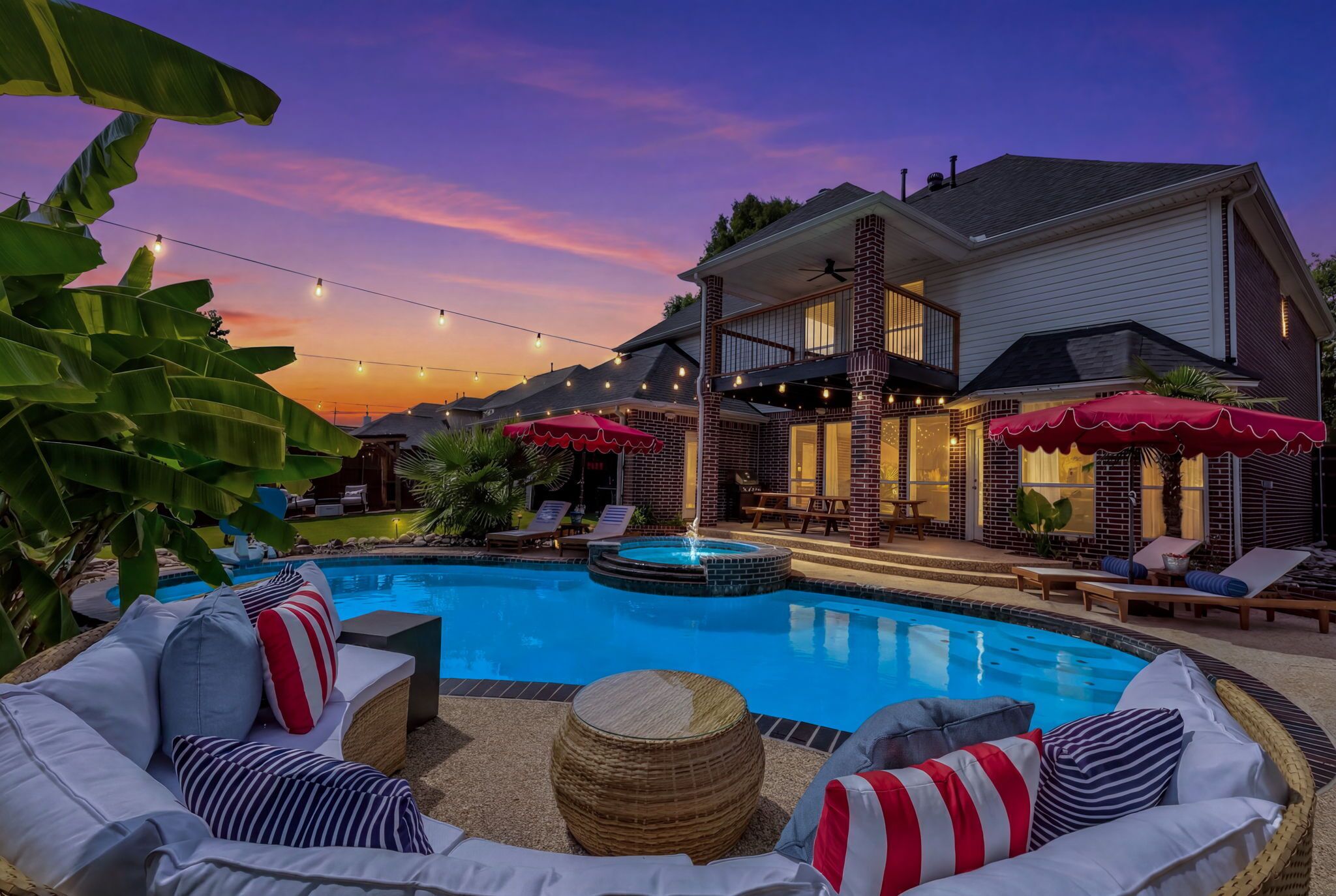 A swimming pool with lounge chairs and a hot tub, surrounded by a house with a balcony and string lights.