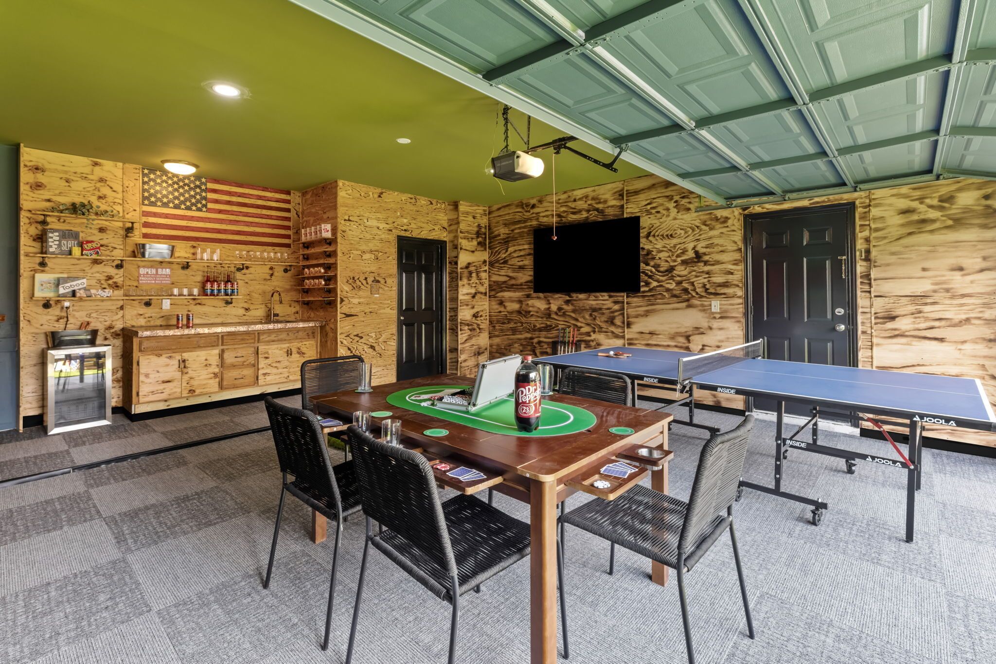 A room with a ping pong table, chairs, a bar area with shelves, and a television mounted on the wall.