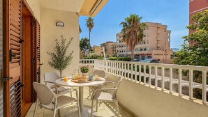 A balcony with a table set for two, chairs, a potted plant, and a view of the street and buildings. - Maison marina Suite - Corfù (Paola)