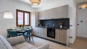 A modern kitchen with wooden cabinets, a black countertop, and a built-in oven. There is a dining area with a table and chairs, and a sofa in the foreground. - Maison marina Suite - Corfù (Paola)