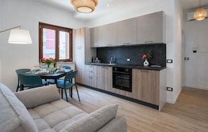 A modern kitchen with wooden cabinets, a black countertop, and a built-in oven. There is a dining area with a table and chairs, and a sofa in the foreground. - Maison marina Suite - Corfù (Paola)
