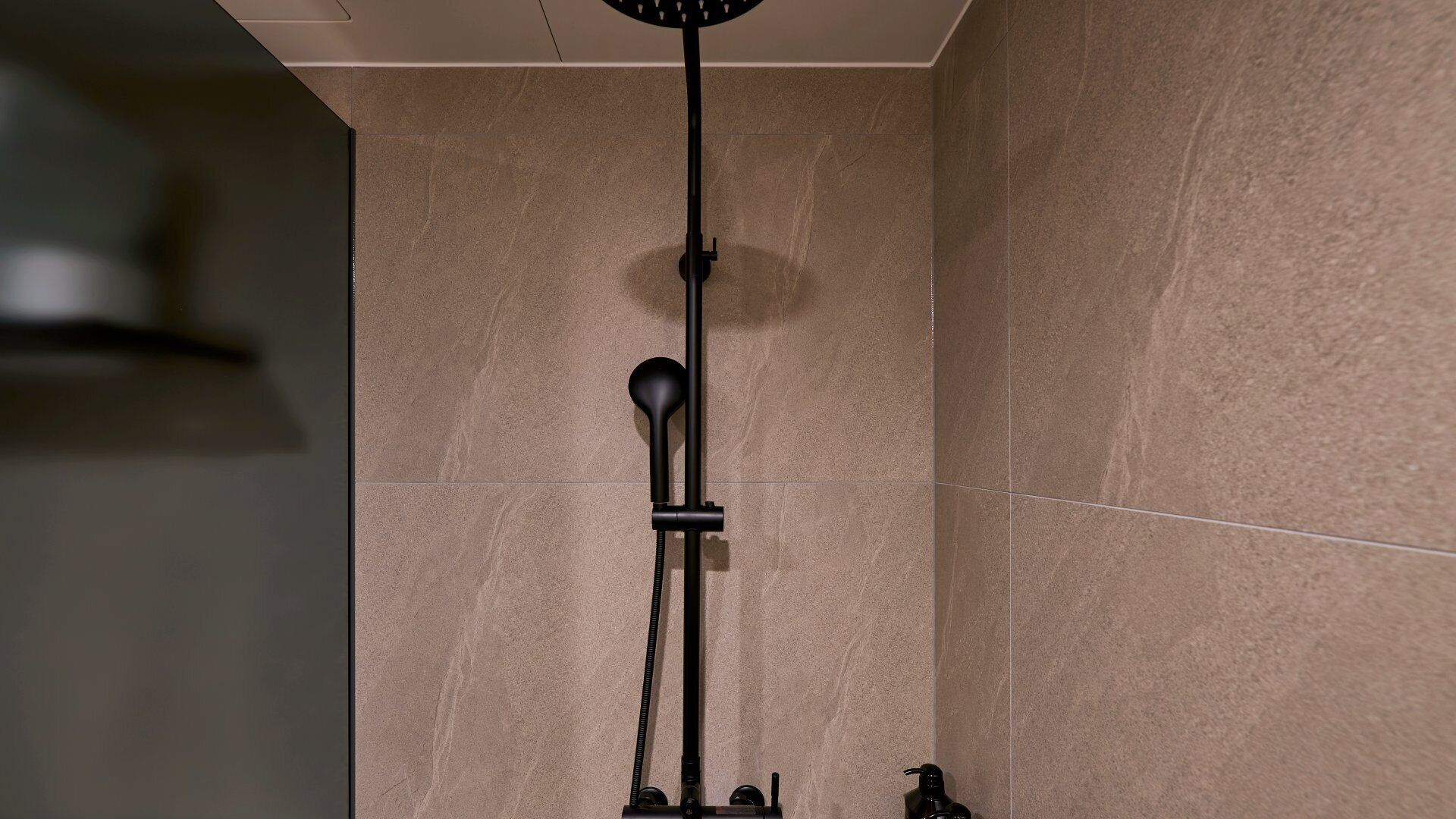 A shower with a black hose and control panel, set against a wall with beige tiles.