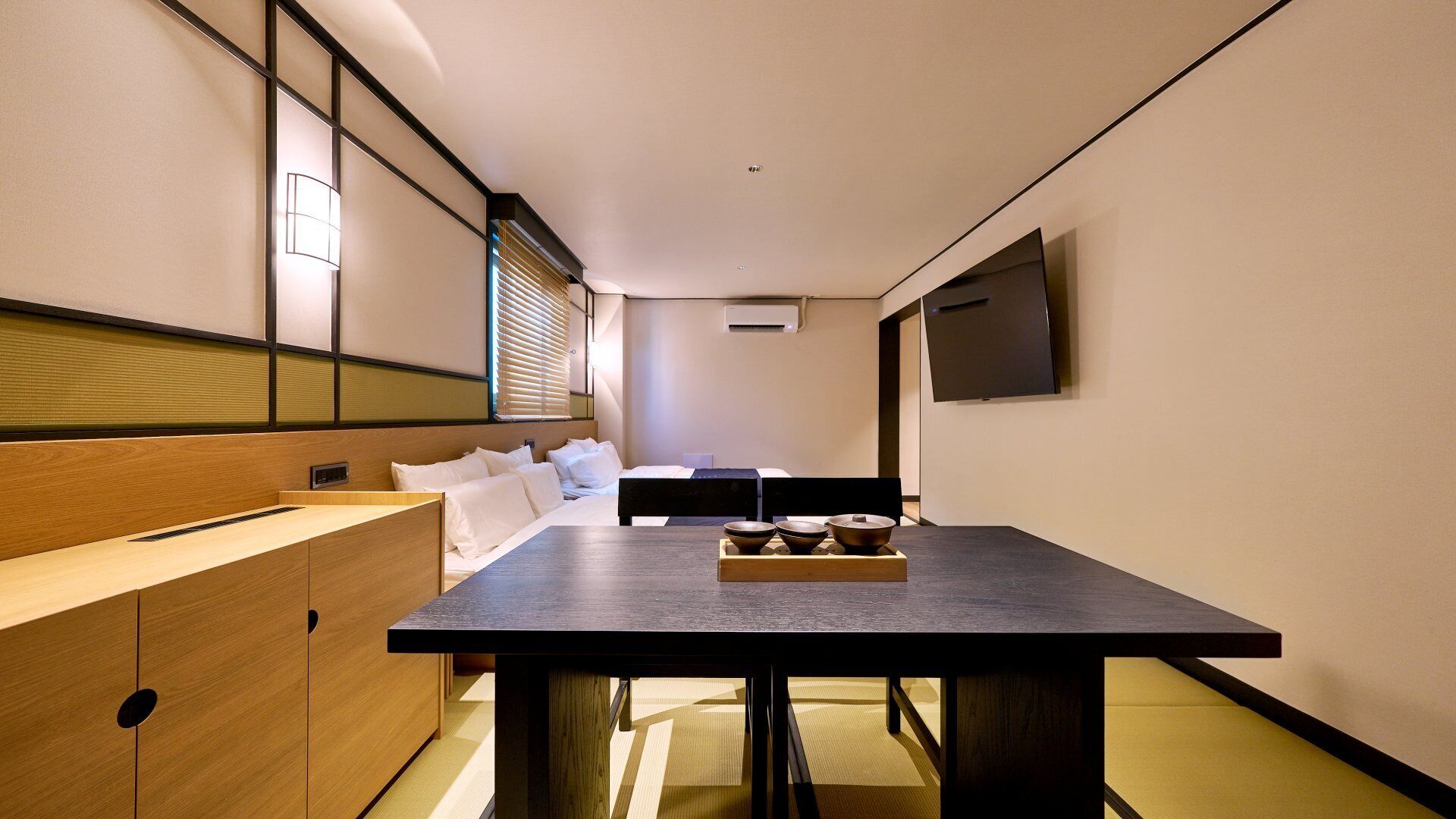 A modern hotel room with a wooden bed, a dining table with a tray of bowls, and a TV mounted on the wall.