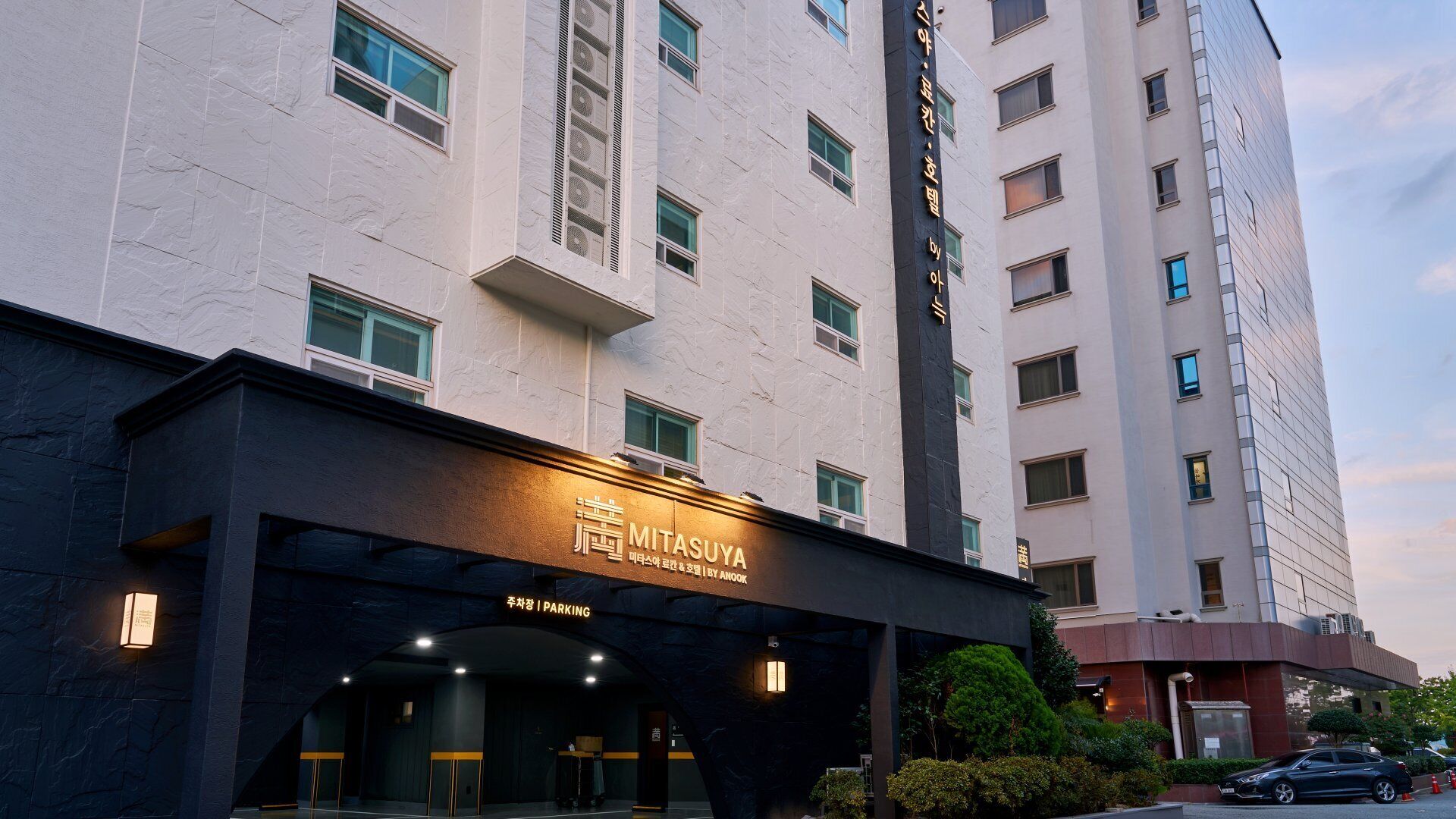 Entrada moderna de un hotel con un cartel que dice 'MITSASUYA' y 'ESTACIONAMIENTO', indicando una zona de aparcamiento.