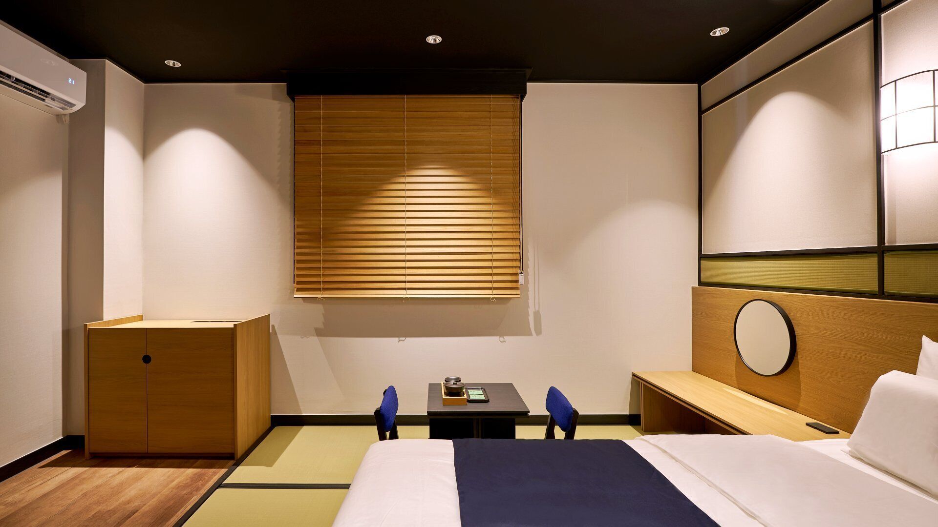 Habitación de hotel con cama, mesita de noche, un escritorio pequeño con silla, armario de madera, espejo montado en la pared y una ventana con persianas.
