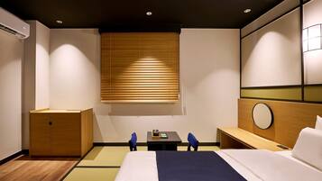 Habitación de hotel con cama, mesita de noche, un escritorio pequeño con silla, armario de madera, espejo montado en la pared y una ventana con persianas.