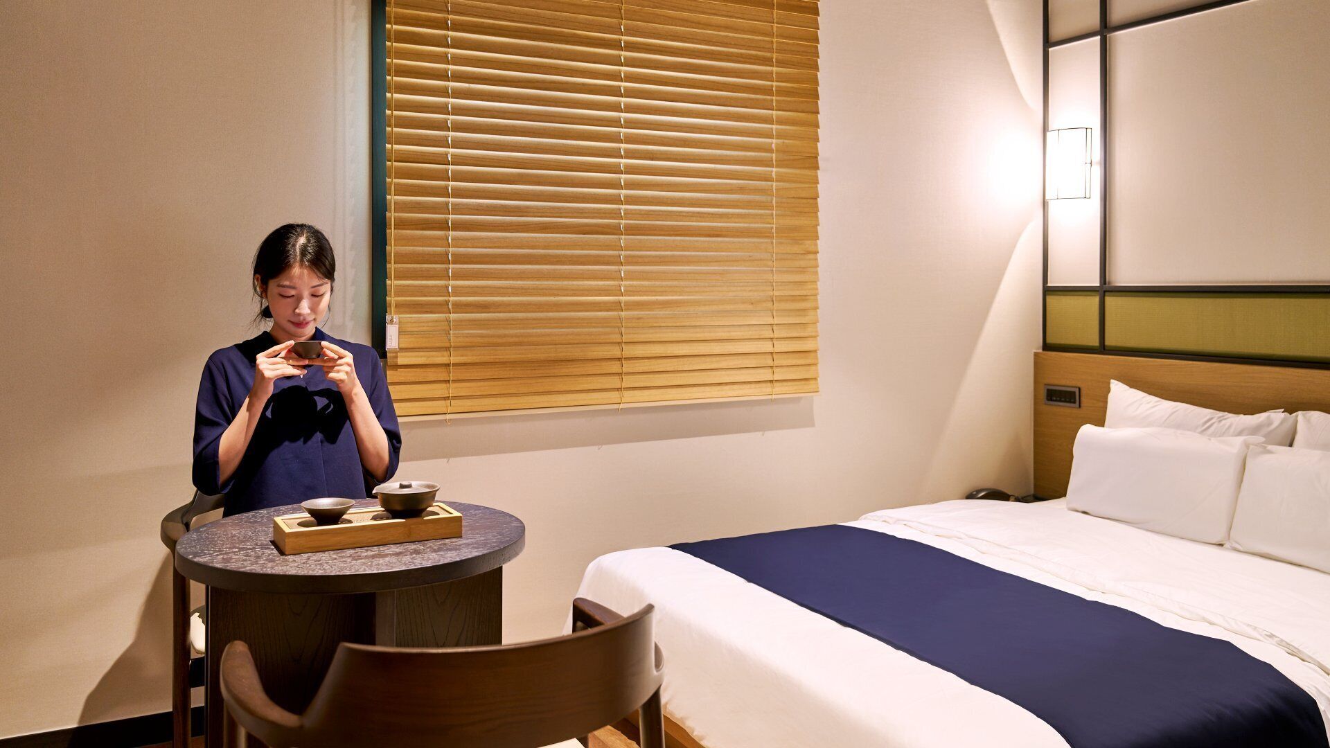 A hotel room with a bed, a table with a bowl and a cup, and a window with blinds.