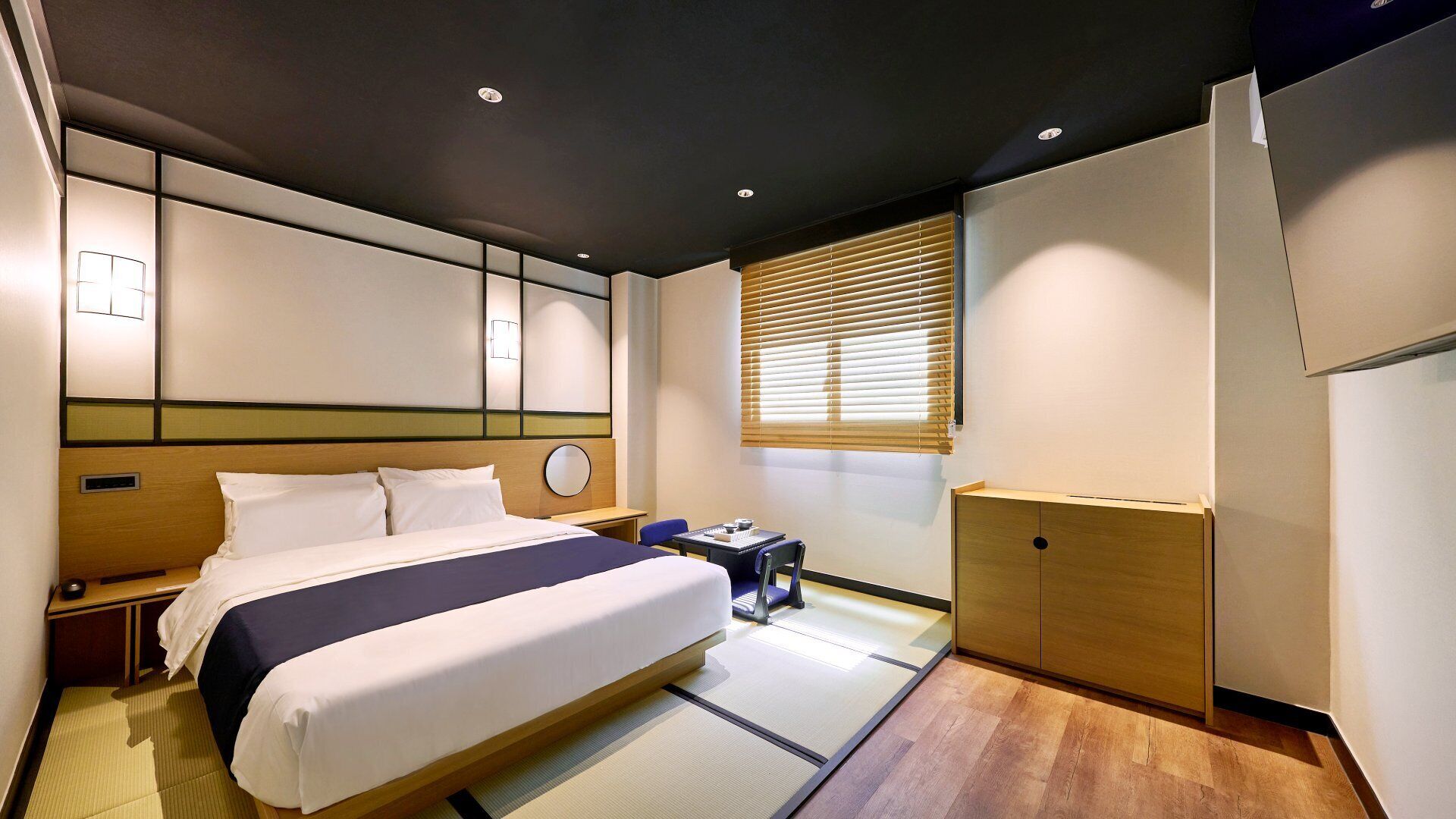 A hotel room with a bed, bedside table, lamp, and a window with blinds.
