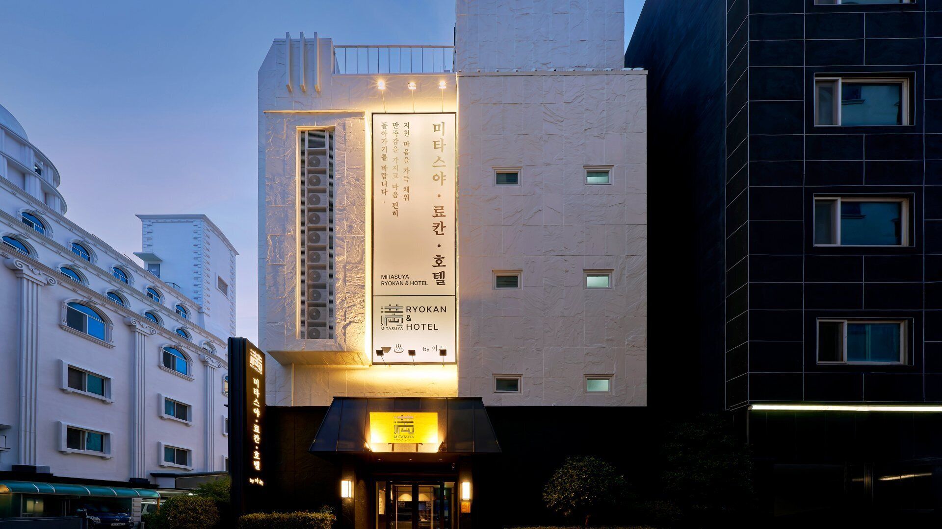 A hotel building with a modern facade, signage indicating 'Ryokan & Hotel', and a well-lit entrance.