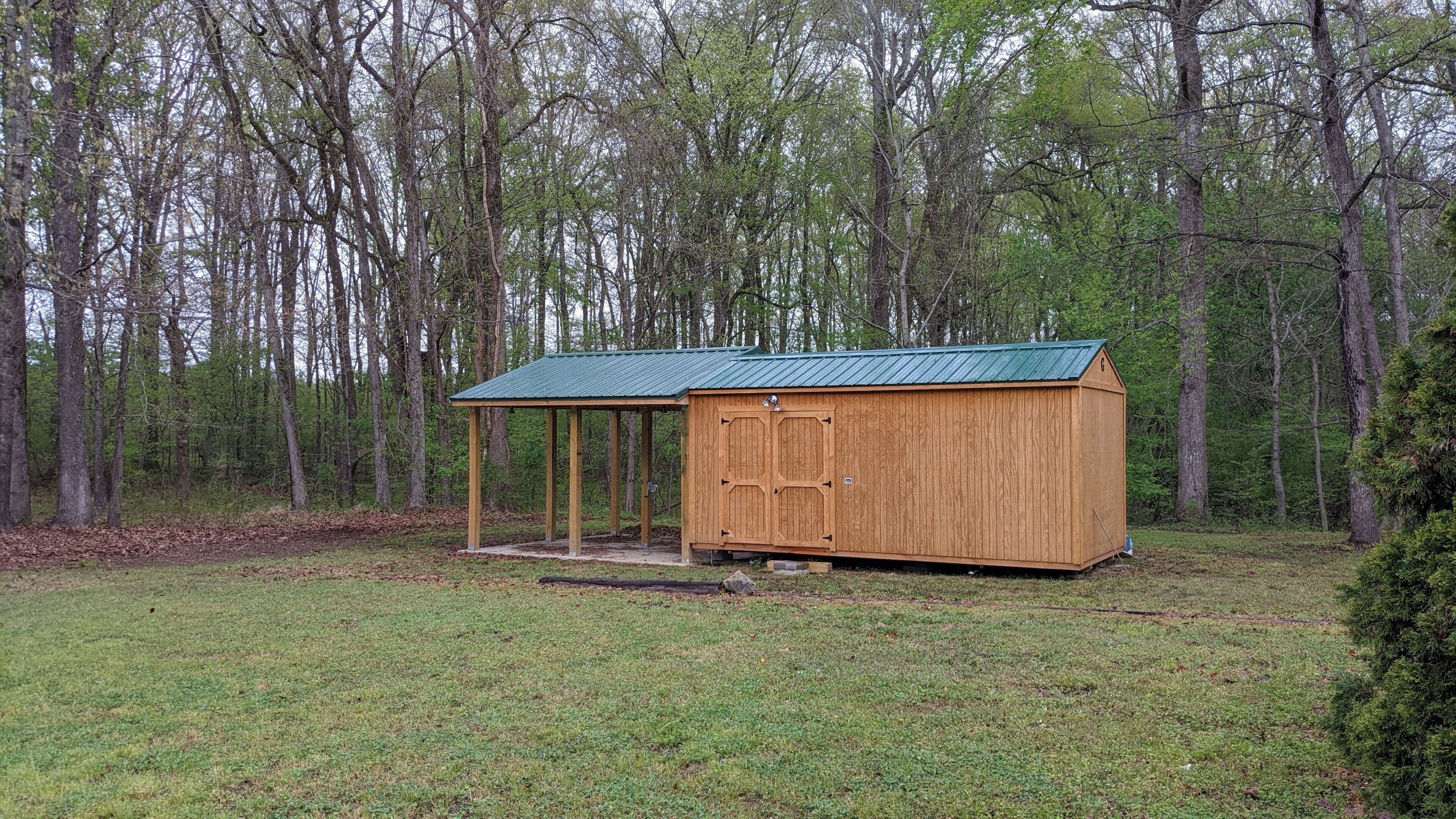 A wooden shed with a green roof in a grassy area surrounded by trees.