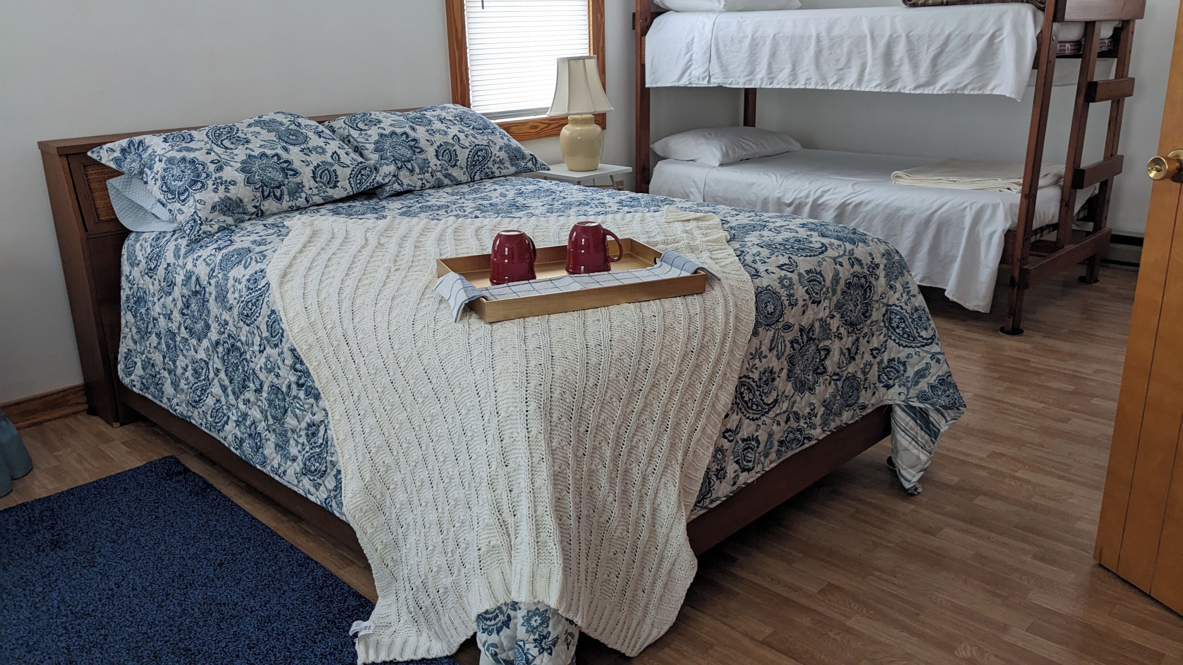 A bed with a patterned bedspread, a knitted throw, and a tray with two red mugs.