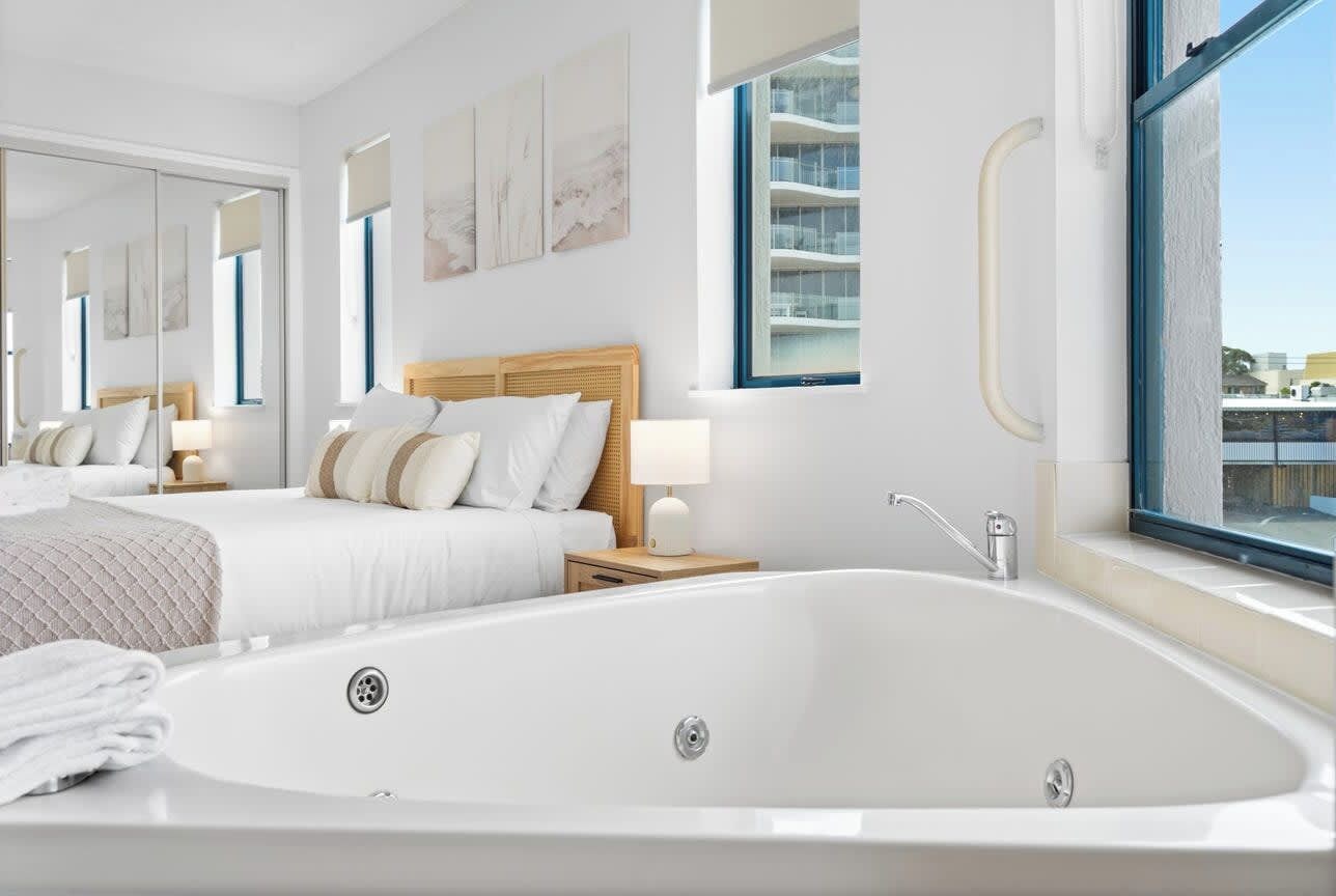 Una habitación de hotel moderna con una bañera grande, una cama con ropa blanca, una mesita de noche con lámpara y vistas a un edificio a través de un ventanal amplio.