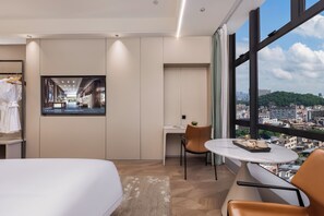 A hotel room with a bed, a desk, a chair, a small table, and a large window offering a city view.