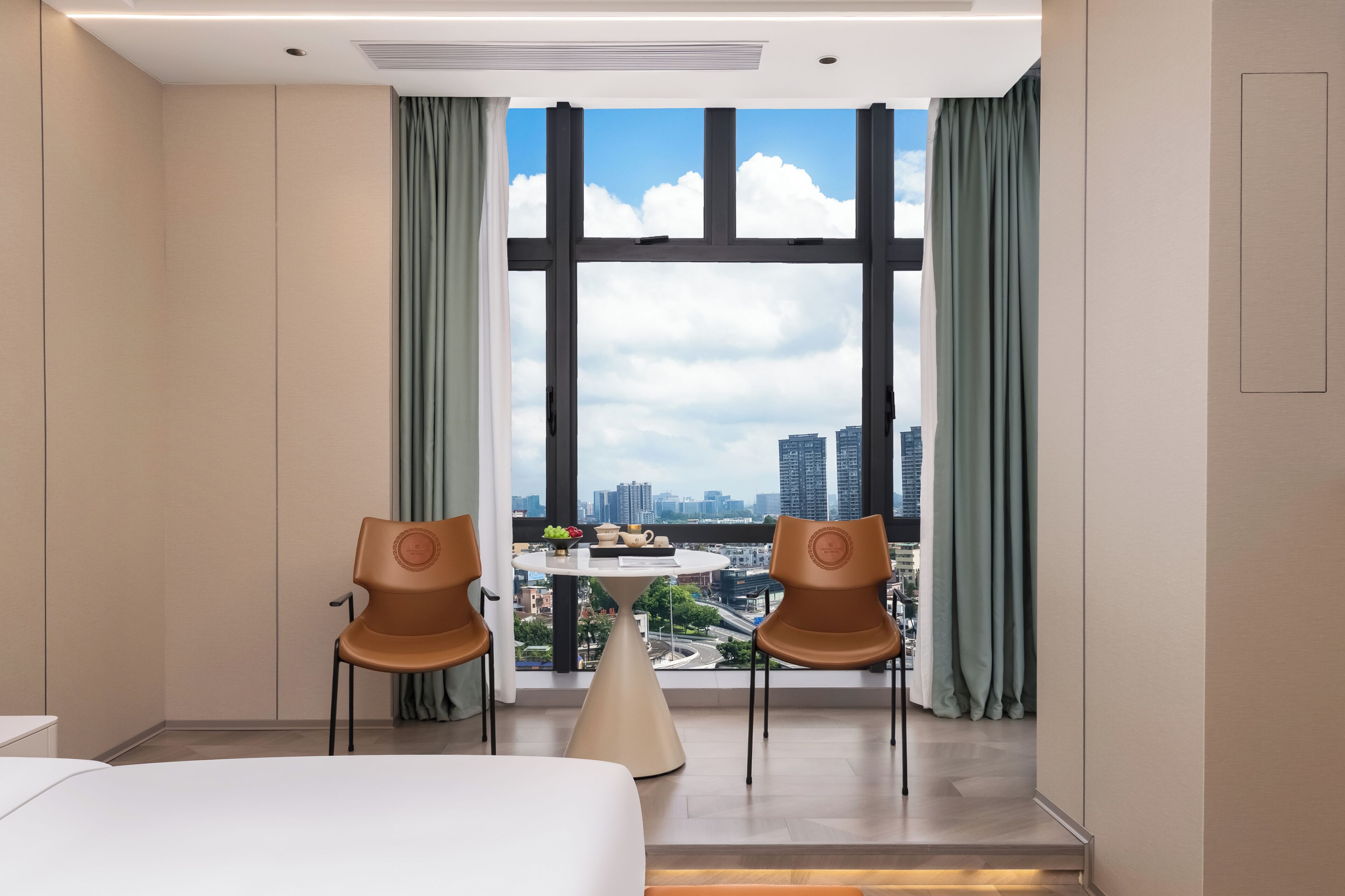 A hotel room with a large window, two chairs, a bed, and a small table with a bowl of fruit.