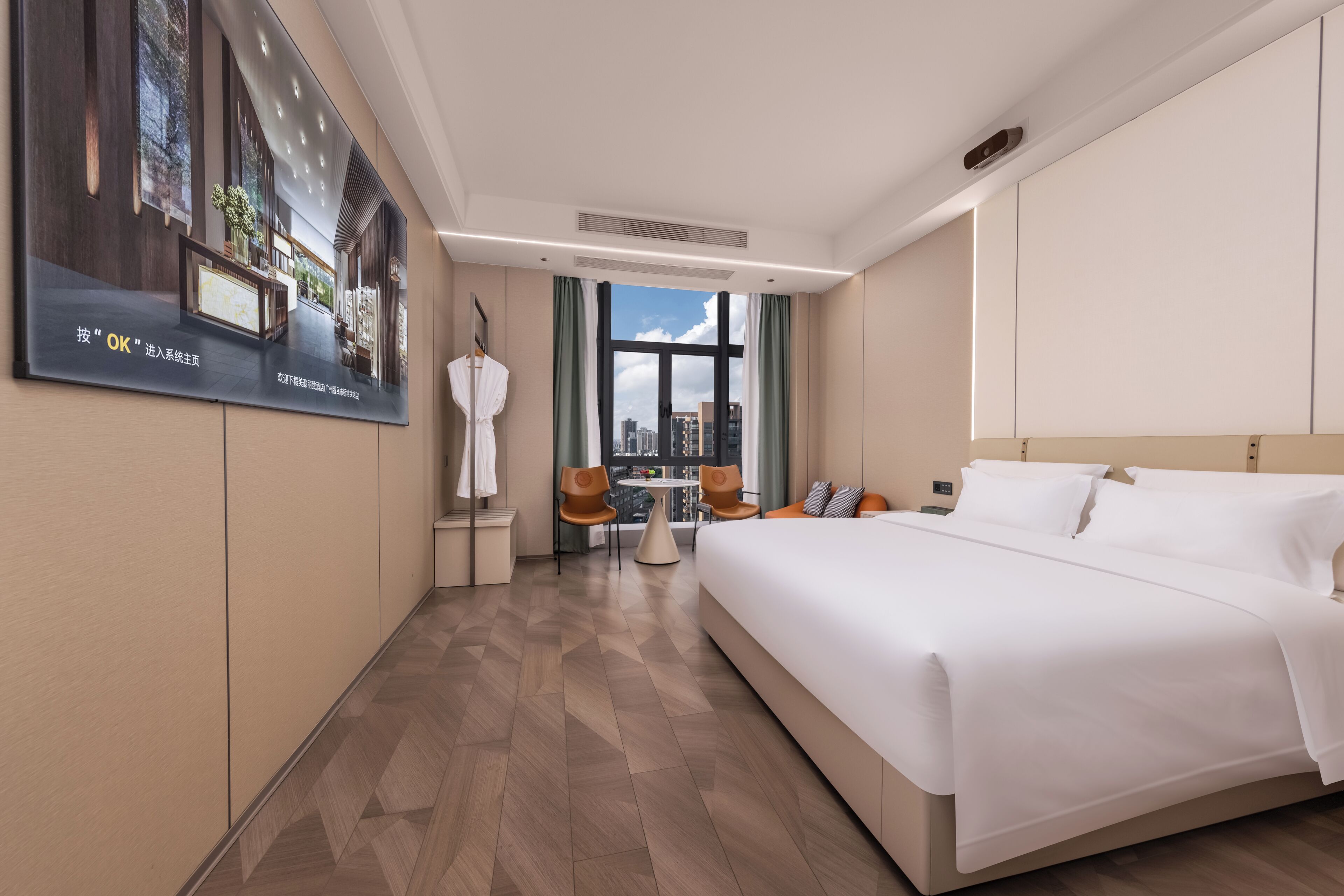 Ein Hotelzimmer mit einem großen Bett, einer Sitzecke mit Tisch und Stühlen, Blick auf die Stadt durch ein großes Fenster und einem Poster an der Wand.