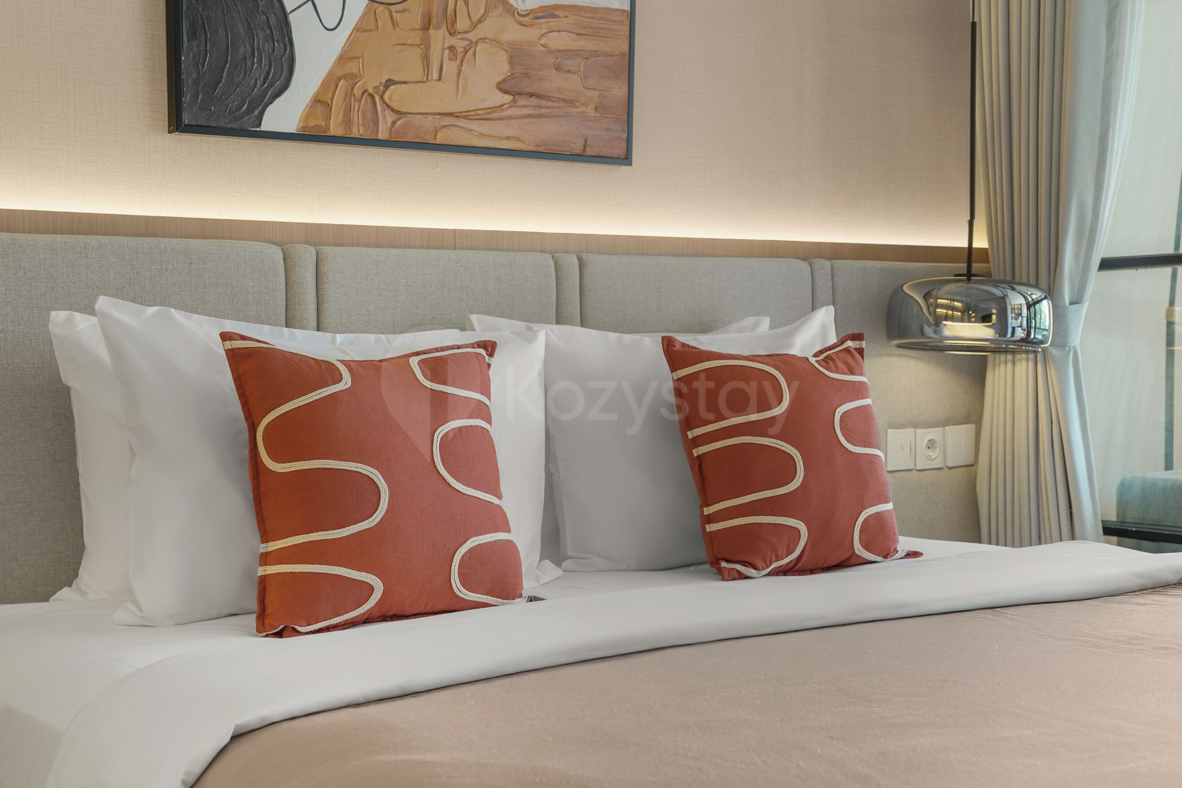 A hotel room with a bed, white and red pillows, a headboard, a painting on the wall, a nightstand with a lamp, and a window with curtains.