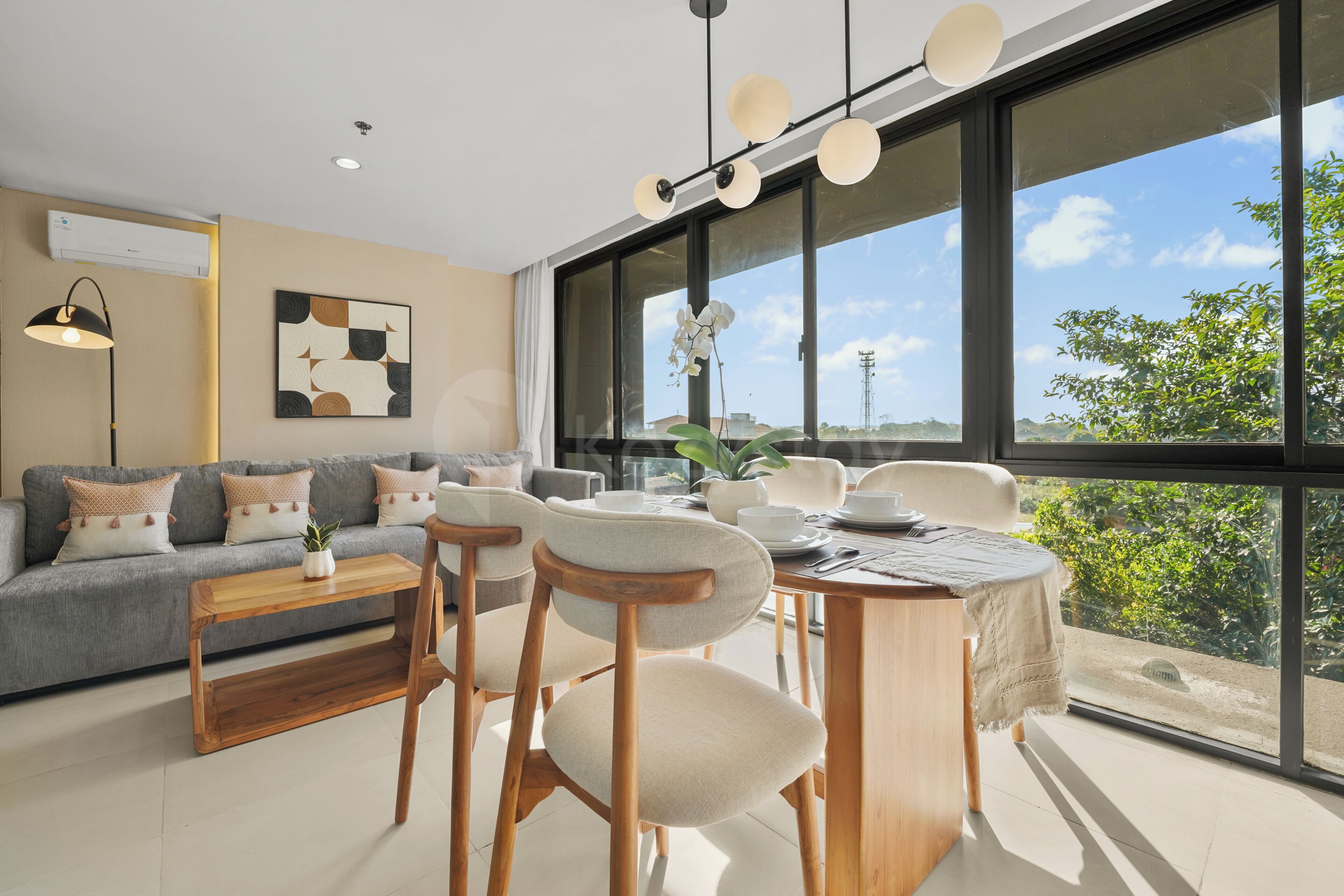 Una sala de estar moderna con un sofá gris, mesa de comedor de madera y sillas, ventanas amplias con vista a follaje.