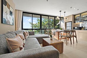 A modern living room with a sofa, a dining area with chairs, a kitchen with a refrigerator, and large windows offering a view of trees. - Jasmine by Kozystay - Nusa Dua (Badung)