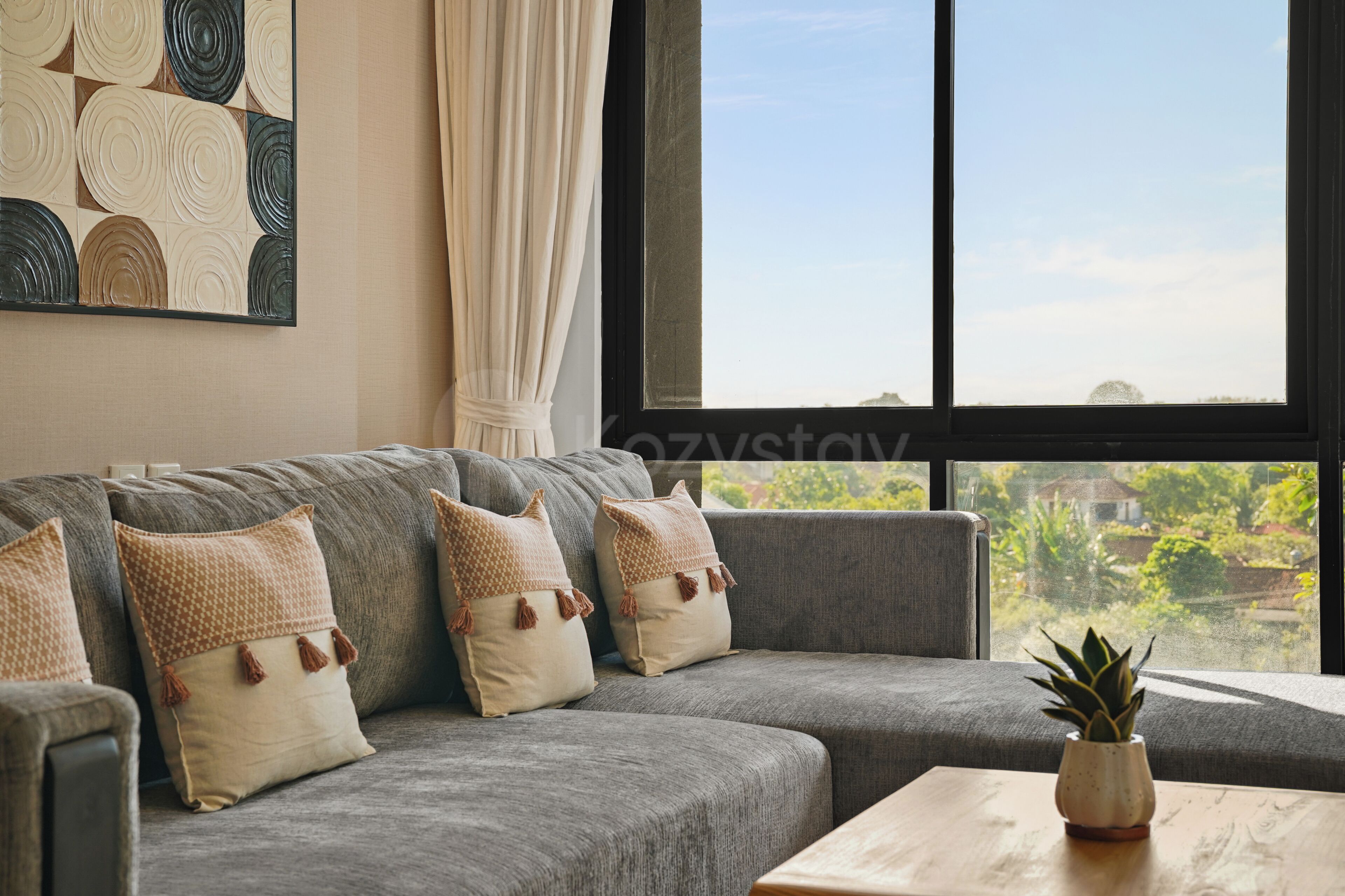 Un sofá gris con cojines estampados, una ventana que ofrece vistas a zonas verdes y una mesa de centro de madera con una planta.