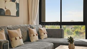 Un sofá gris con cojines estampados, una ventana que ofrece vistas a zonas verdes y una mesa de centro de madera con una planta.