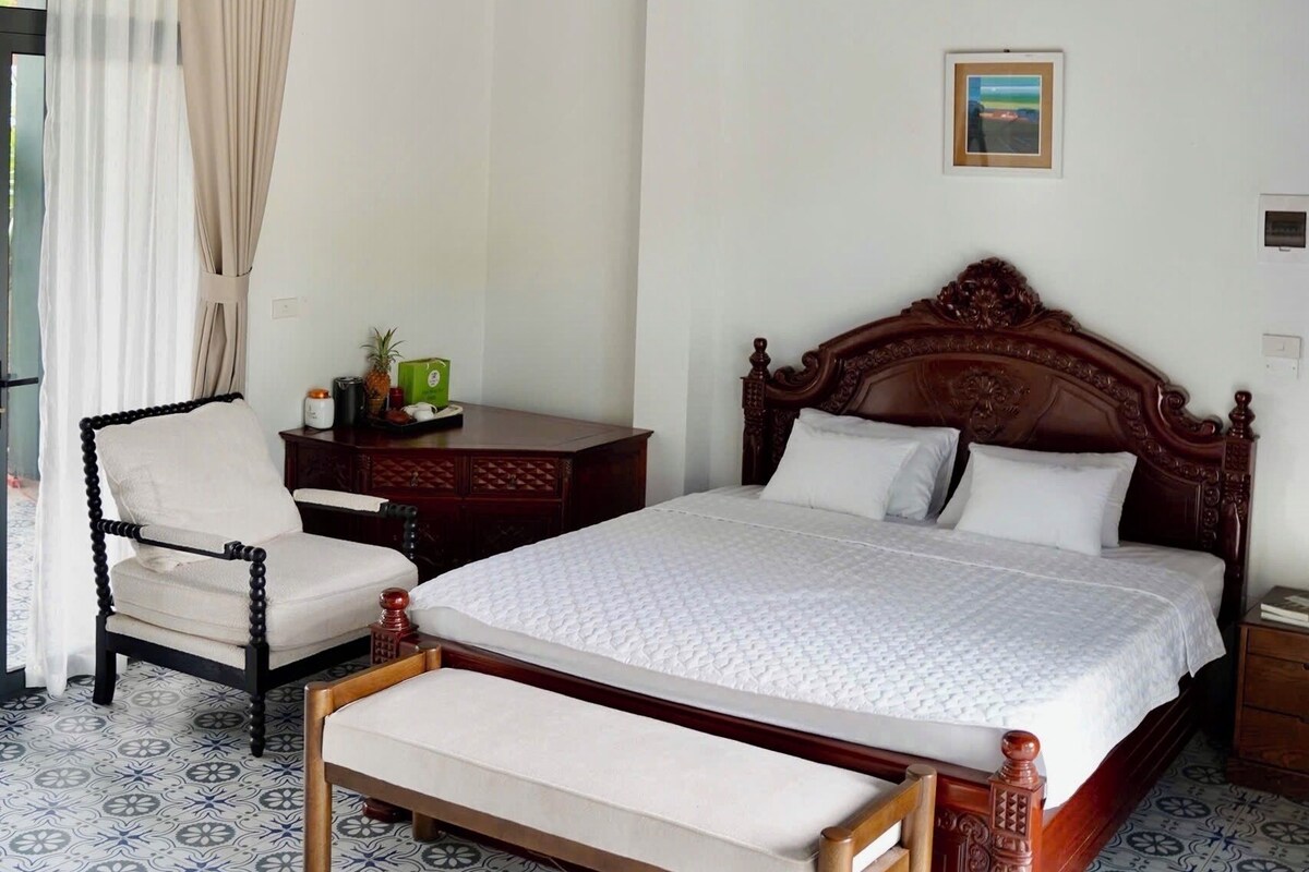 A bedroom with a bed, a chair, a bedside table, a hanging light, and a painting on the wall.