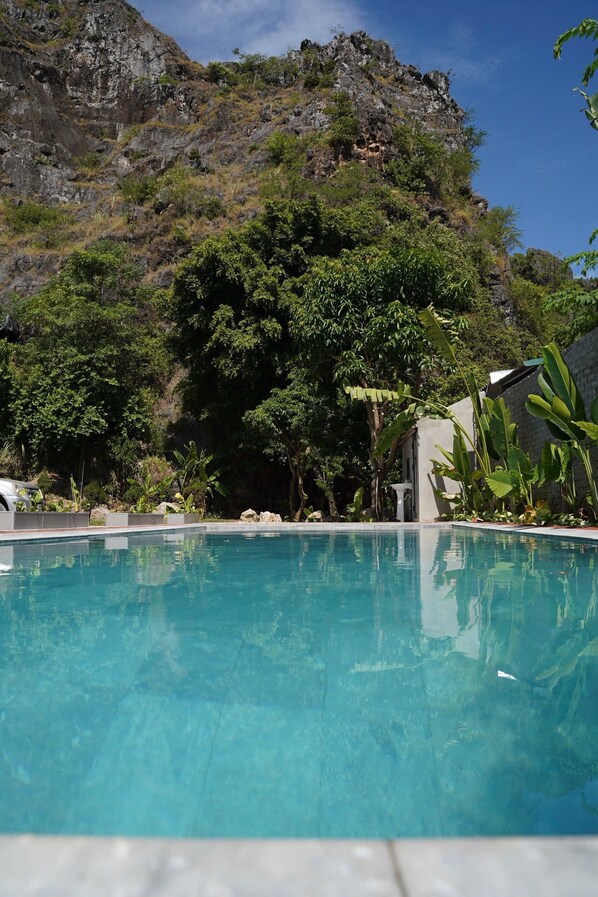 A swimming pool with clear blue water, surrounded by lush greenery and a rocky hillside in the background.