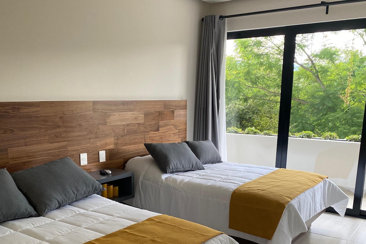 A hotel room with two beds, a wooden headboard, a nightstand, and a view of greenery outside.