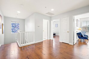 A spacious hallway with wooden flooring, white walls, and a staircase with a white railing. There are doors leading to other rooms, and a blue chair is visible in the foreground.