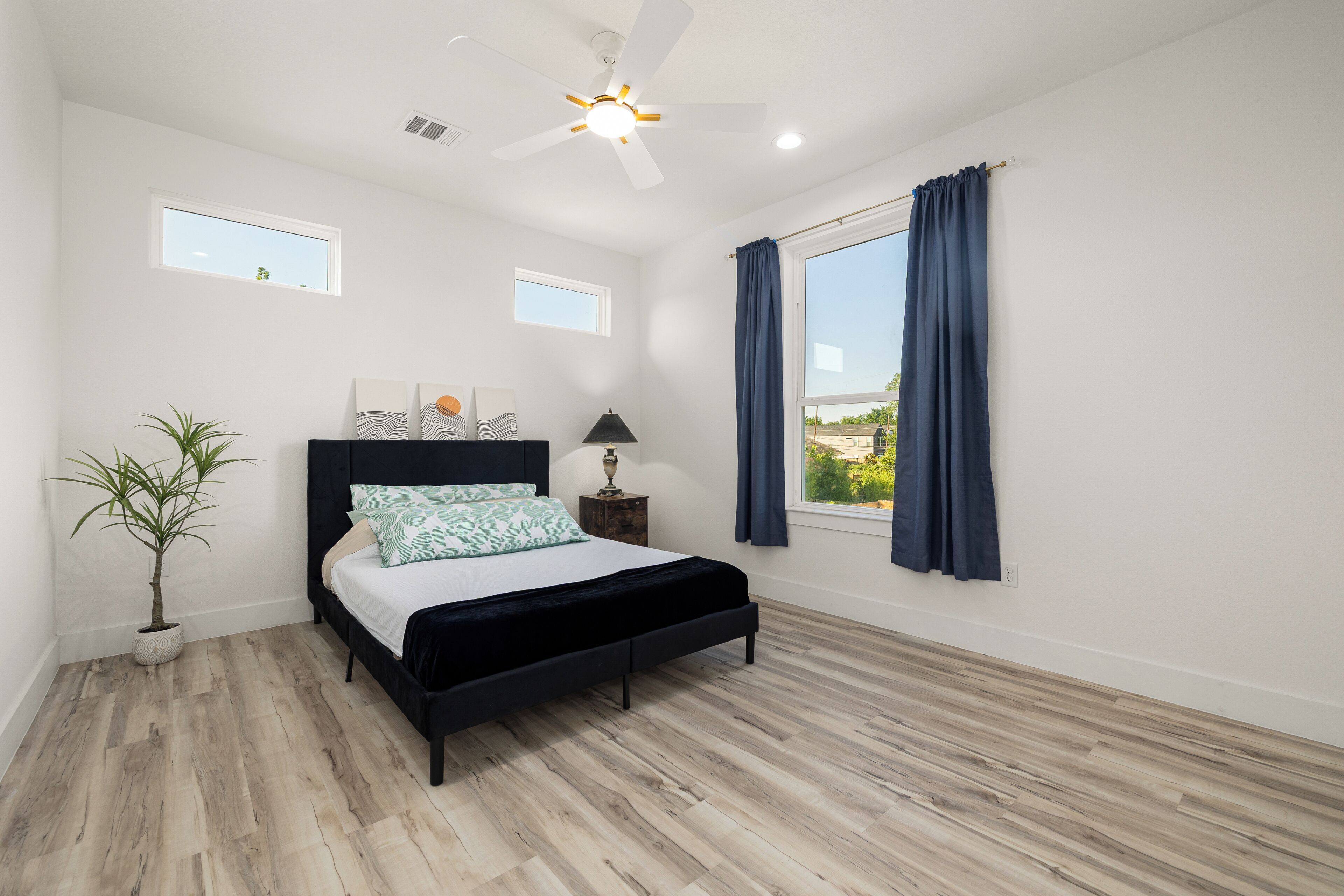 Quarto com cama, mesa de cabeceira, abajur, ventilador de teto e janela com cortinas.