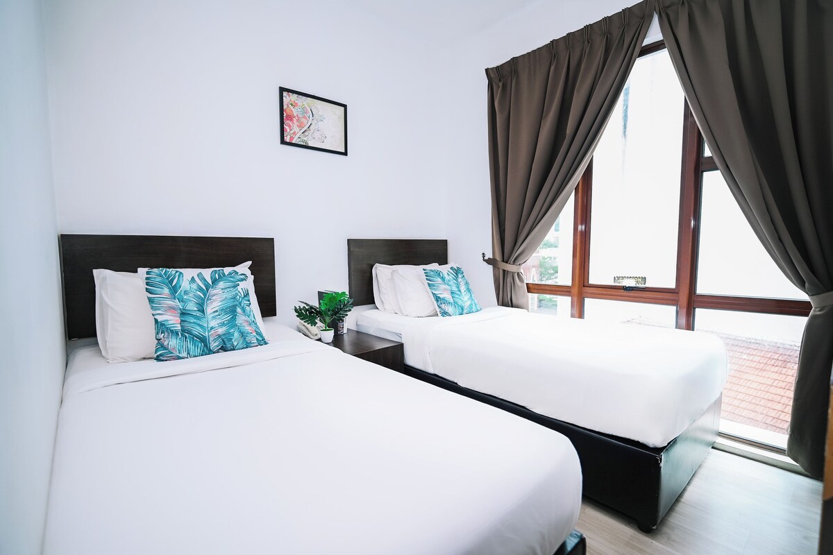 A hotel room with two beds, a small table with a plant, a framed picture on the wall, and a window with curtains.