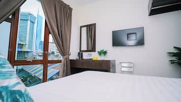 A hotel room with a bed, a desk with a mirror, a flat-screen TV, and a view of a cityscape through a large window.