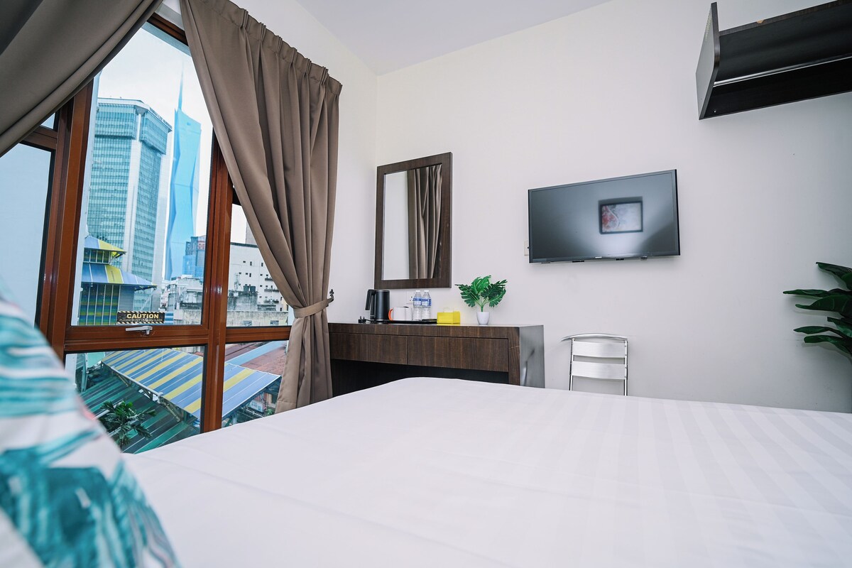 Habitación de hotel con cama, escritorio con espejo, televisor de pantalla plana y vista a un paisaje urbano a través de un ventanal grande.