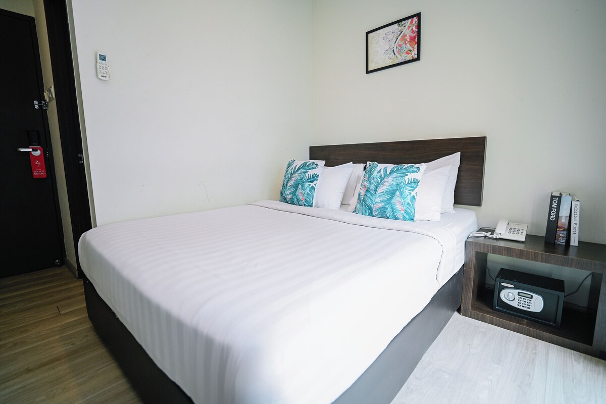 A hotel room with a bed, a bedside table with a phone, and a wall-mounted thermostat.