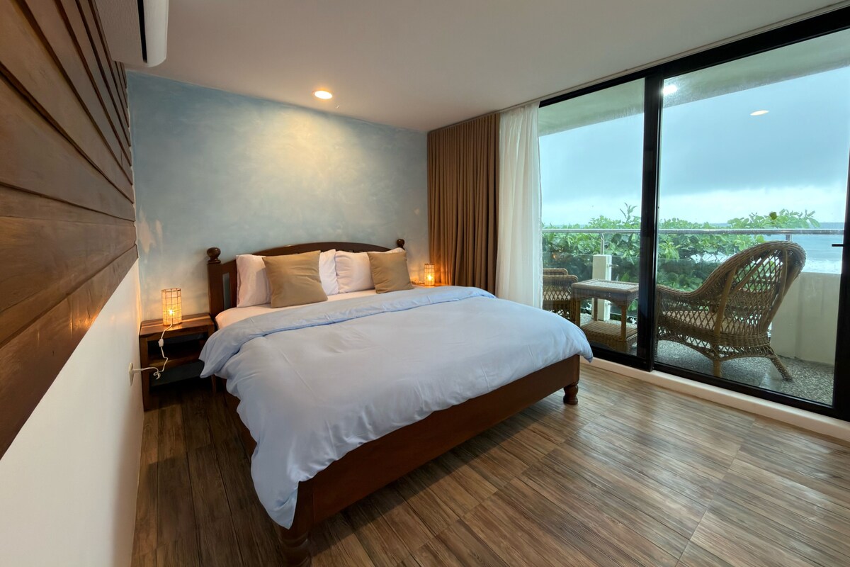 Une chambre avec un grand lit, une table de chevet avec une lampe, une fenêtre donnant sur un paysage verdoyant et un balcon avec une chaise.